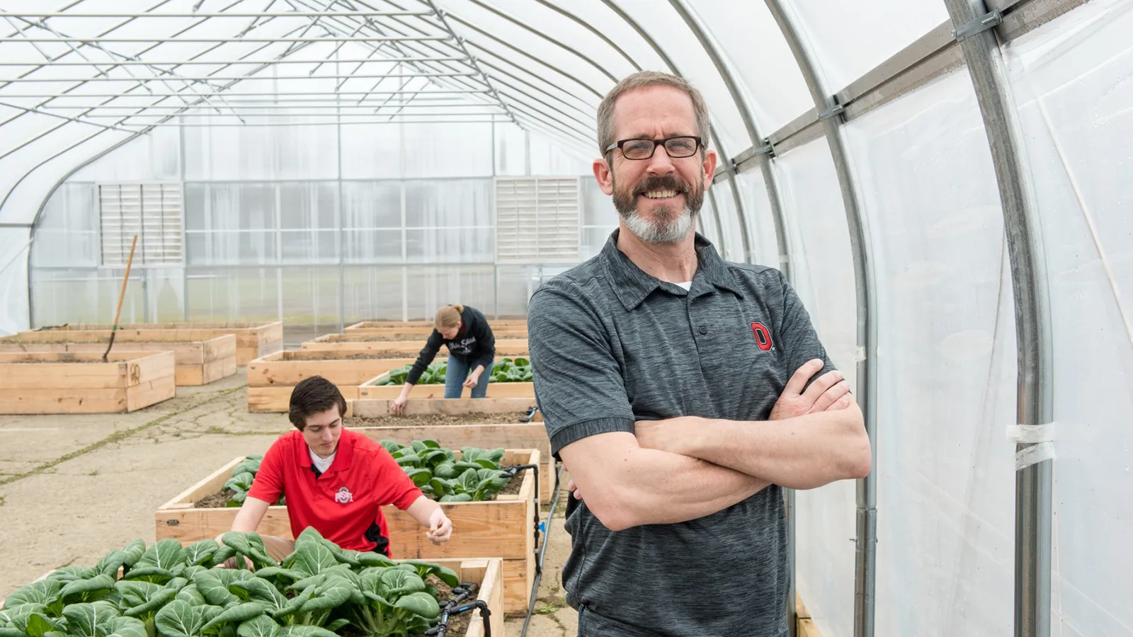 Kip Curtis standing in a green house as students tend plants int he background