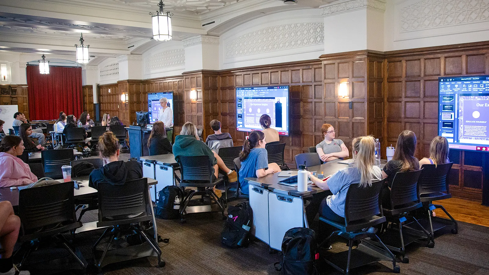 Students sitting around collaboration tables as a faculty member tecahes from the front of the class